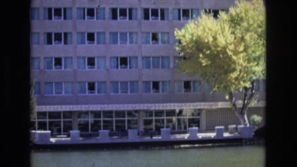 Closer 1974 Colorado Springs home-movie view of the building's repetitive window grid, waterside terrace, calm water, and a large yellow-green autumn tree at the right edge.