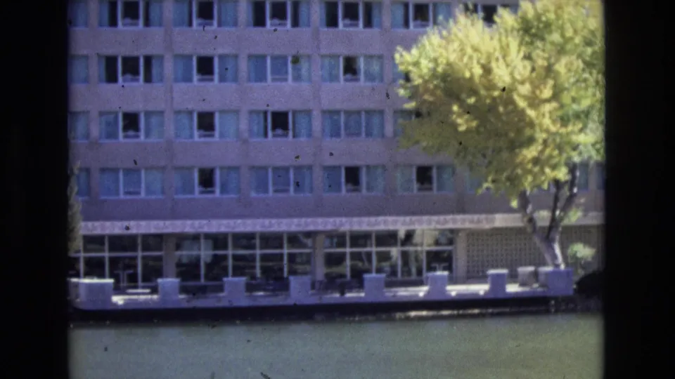 1974 Colorado Springs frame showing the multi-story facade across calm water, with a large yellow-green tree overlapping the right side and a ground-level terrace beneath the windows.