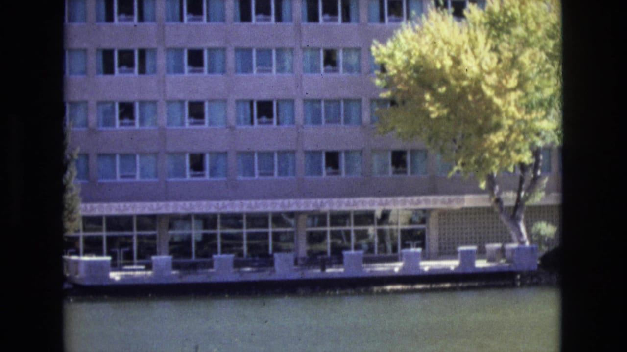 1974 Colorado Springs home-movie frame showing a multi-story waterside building, rows of windows, a glassy ground floor, calm water, and a yellow-green tree in bright sun.