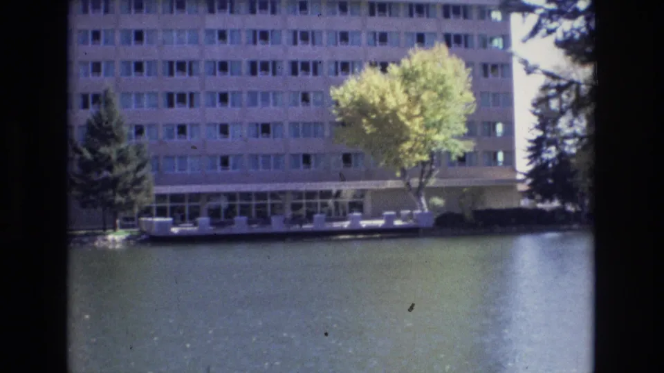 Wider 1974 Colorado Springs home-movie frame showing a waterside building, a yellow-green tree, evergreen trees, a pale waterfront edge, and calm reflective water.