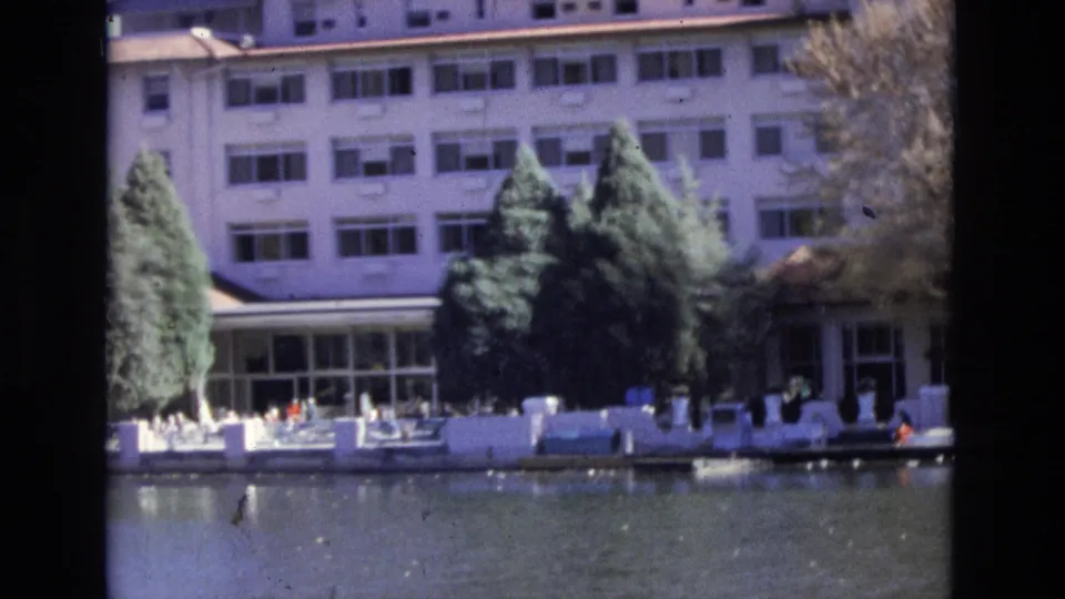 1974 Colorado Springs frame showing a waterside terrace below a multi-story building, evergreen trees, outdoor seating, calm water, and small distant people near the waterfront.
