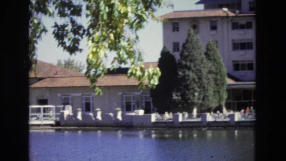 1974 Colorado Springs 8mm frame looking across water toward lower hotel structures, leafy tree branches, white railings, and tall evergreens.
