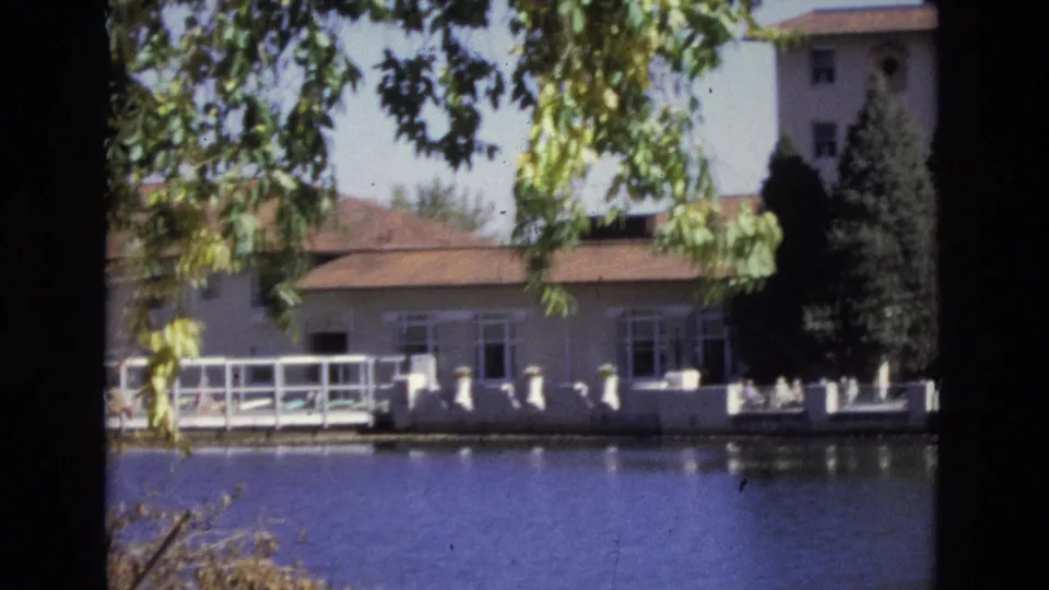 1974 Colorado Springs 8mm frame showing the lakeside edge of the hotel property, with white railings, a low red-roofed building, trees, and calm water.