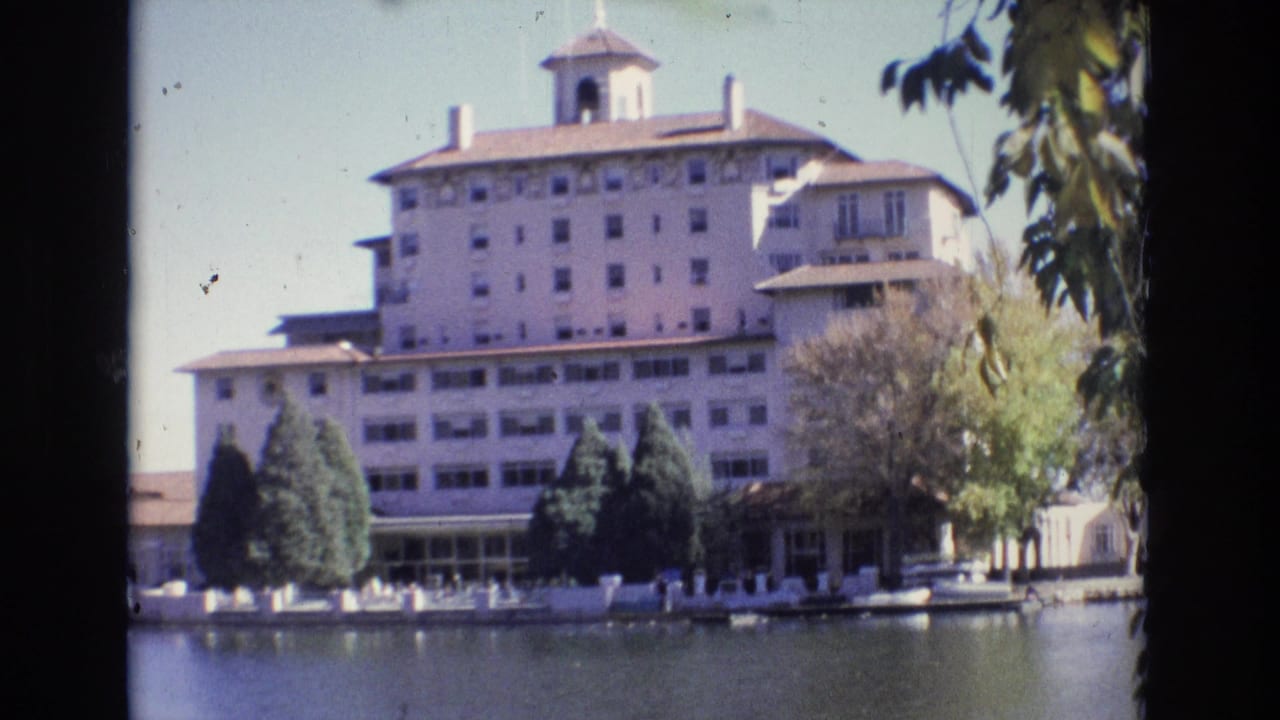 Hotel facade across a lake with trees and a white waterfront fence.