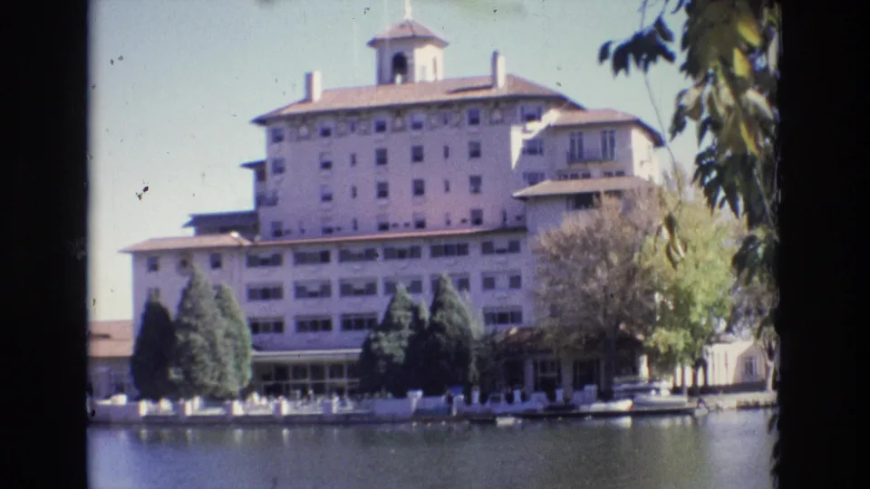 1974 8mm view of the Broadmoor Hotel in Colorado Springs seen across calm water, with evergreens, a white fence, and distant visitors along the shoreline.