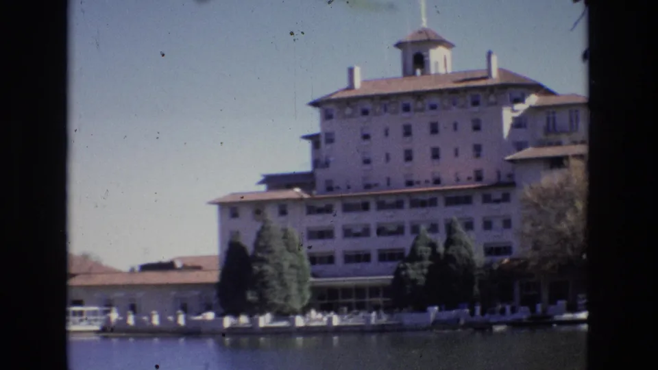 Soft 1974 8mm frame of the Broadmoor Hotel in Colorado Springs with the lake in the foreground, dark trees, and a low waterfront fence.
