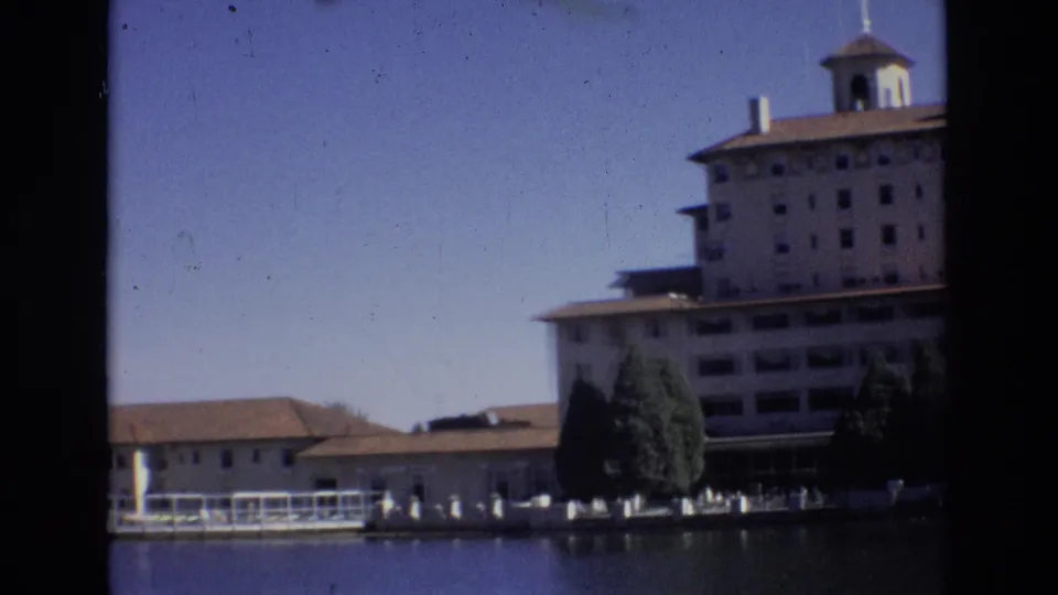 Later frame from the 1974 Colorado Springs clip showing a cropped lakeside view of the Broadmoor Hotel, blue-tinted water, white fence, and trees.