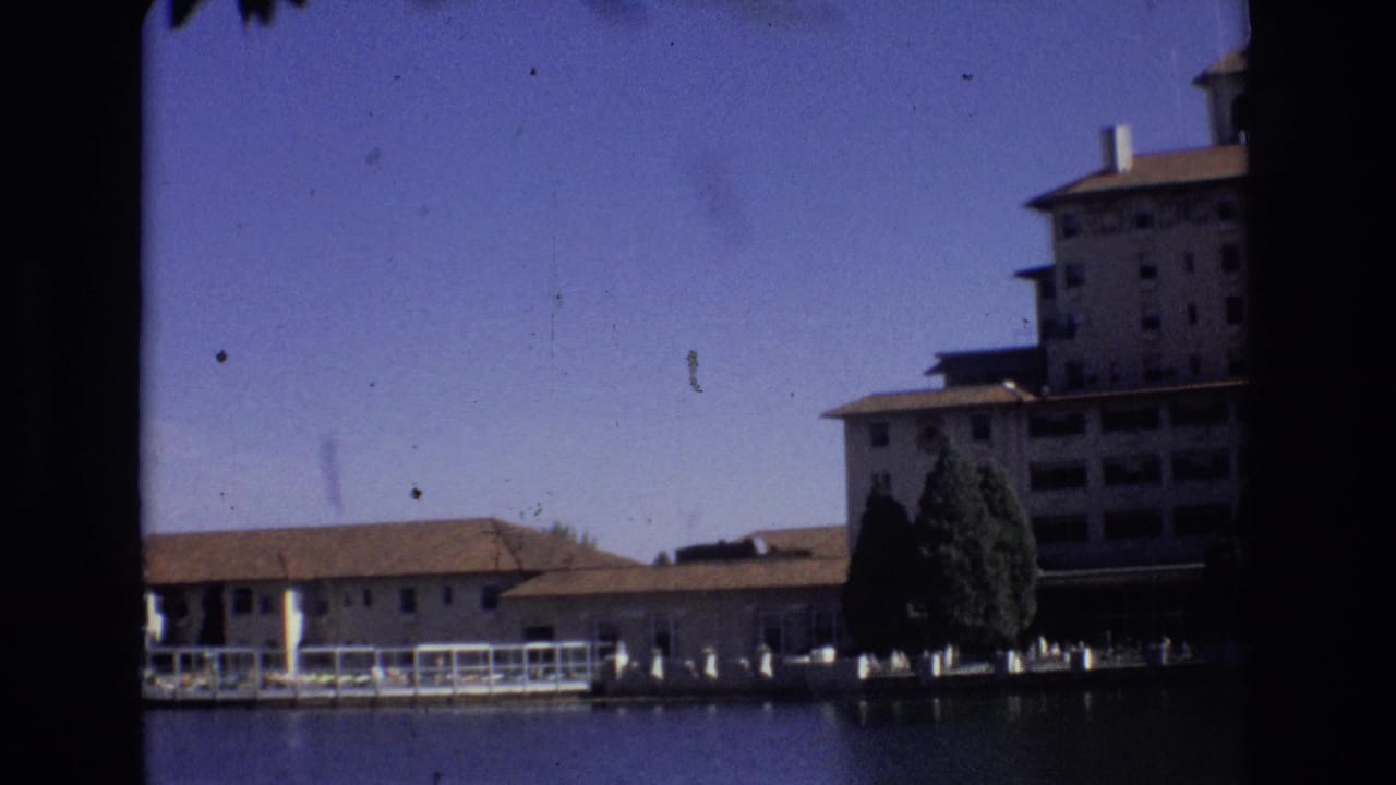 1974 Colorado Springs home-movie frame showing a lakeside resort complex, including a tall building, lower waterfront structures, dock railings, trees, and open sky.