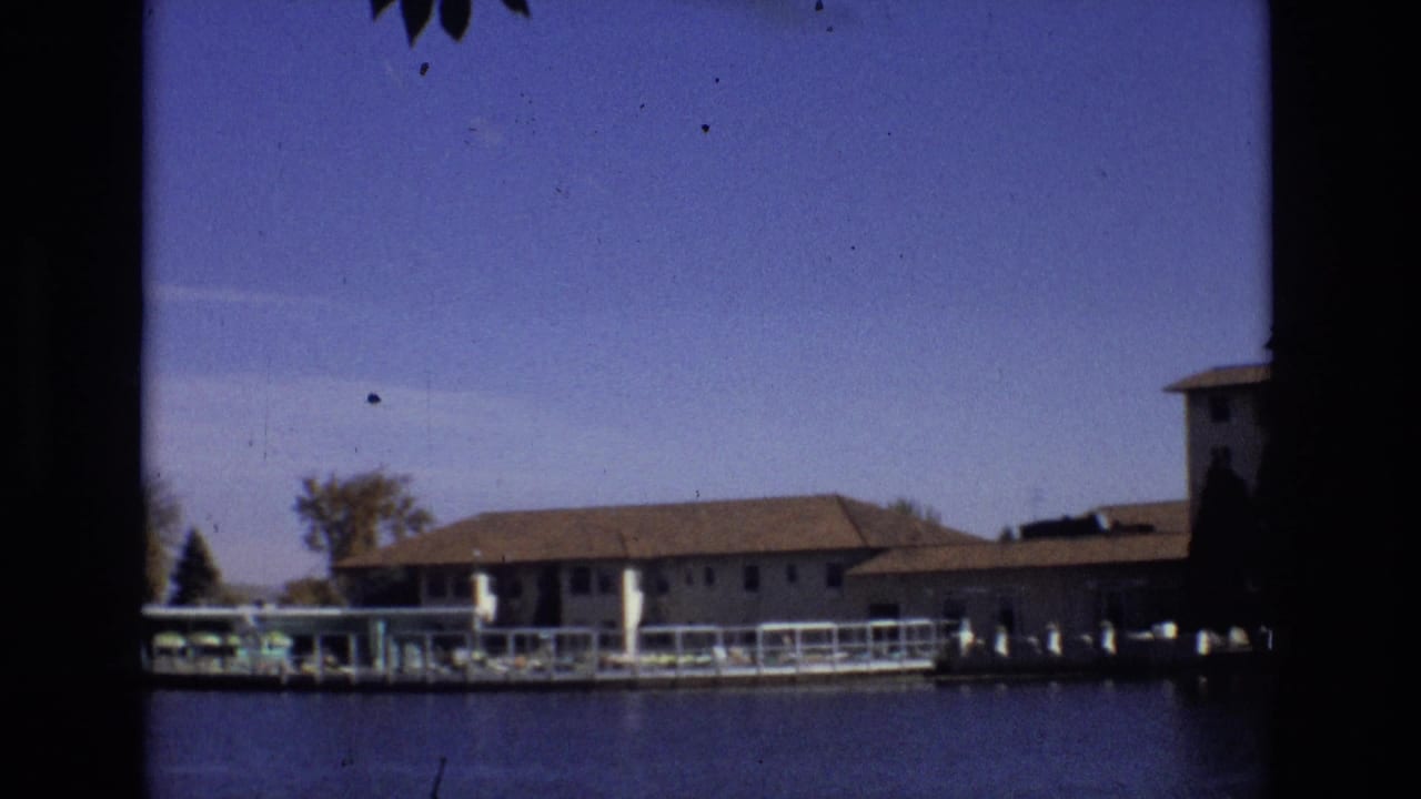 1974 Colorado Springs 8mm frame with a low resort building along the shoreline, dock structures, trees, water, and a partial building at the right edge.