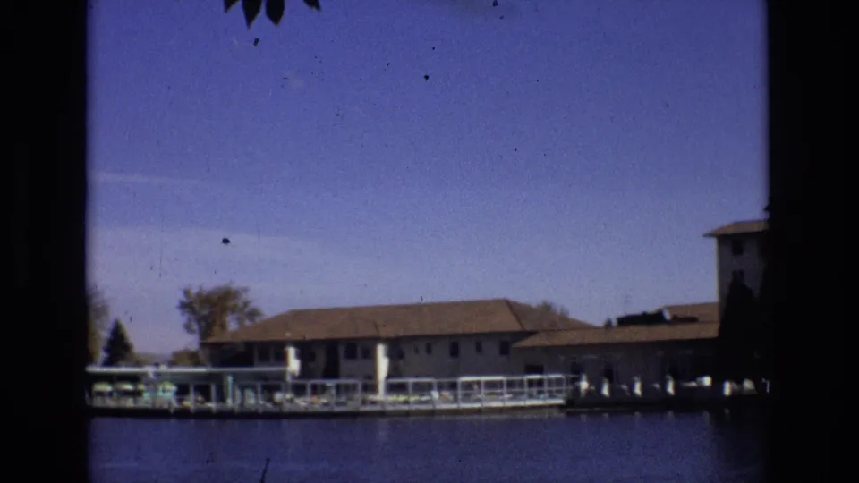 1974 Colorado Springs 8mm frame with a low resort building along the shoreline, dock structures, trees, water, and a partial building at the right edge.