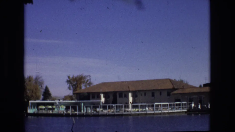 1974 Colorado Springs home-movie frame showing a low lakeside building behind dock railings, with trees at the shoreline and water in the foreground.