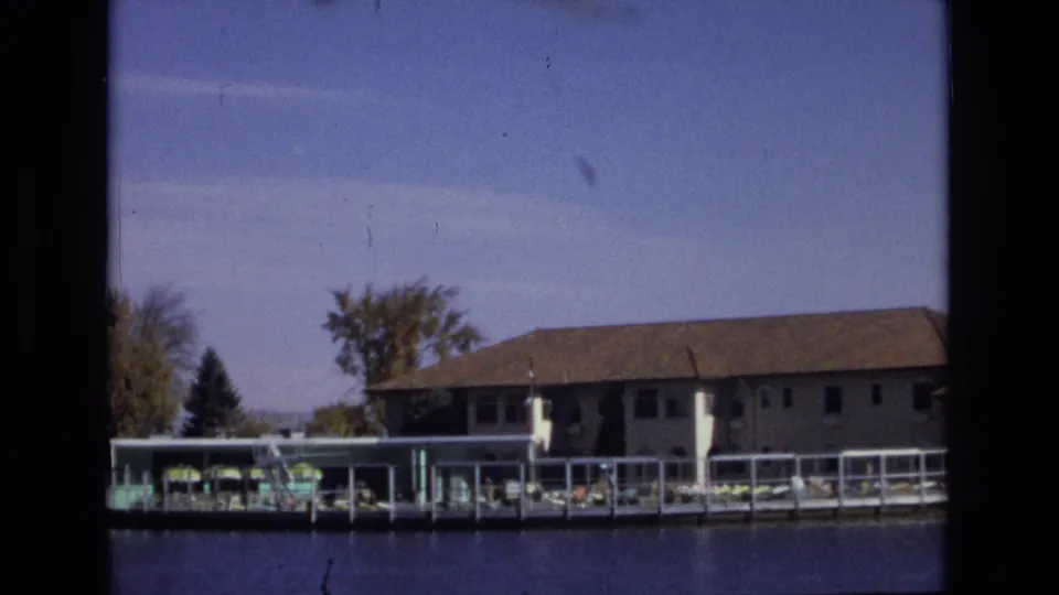 1974 Colorado Springs 8mm frame showing a lakefront resort building, dock railings, trees, still water, and clear sky with visible film grain and gate edges.