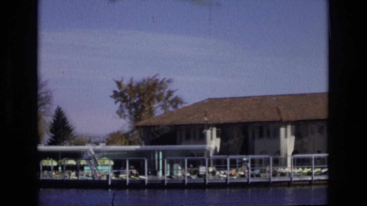1974 Colorado Springs home-movie frame showing a lakeside hotel pool deck with green umbrellas, lounge chairs, white walls, brown roof, trees, and blue water in the foreground.