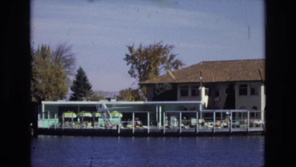 1974 Colorado Springs 8mm frame of a lakeside hotel recreation deck, with green umbrellas, rows of lounge chairs, a white building, trees, and calm blue water.