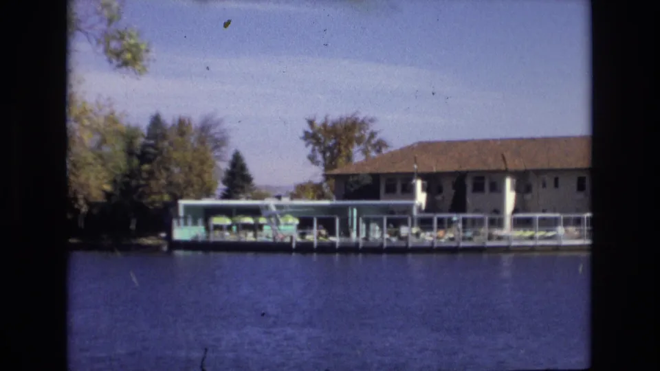 Wider 1974 Colorado Springs home-movie view across blue water toward a white lakeside hotel building, pool deck, green umbrellas, lounge chairs, and shoreline trees.