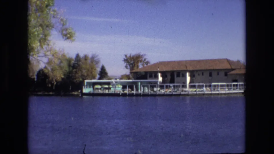 1974 Colorado Springs frame showing the hotel pool deck from farther back, with blue water filling the foreground, trees along the shore, green umbrellas, and the brown-roofed building behind.
