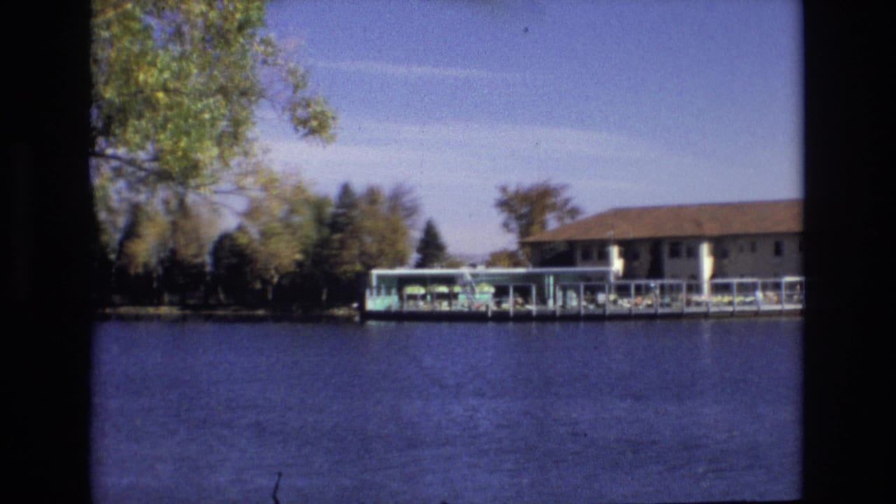 1974 Colorado Springs 8mm frame showing a long low lakeside building and covered dock across blue water, with shoreline trees and clear sky.
