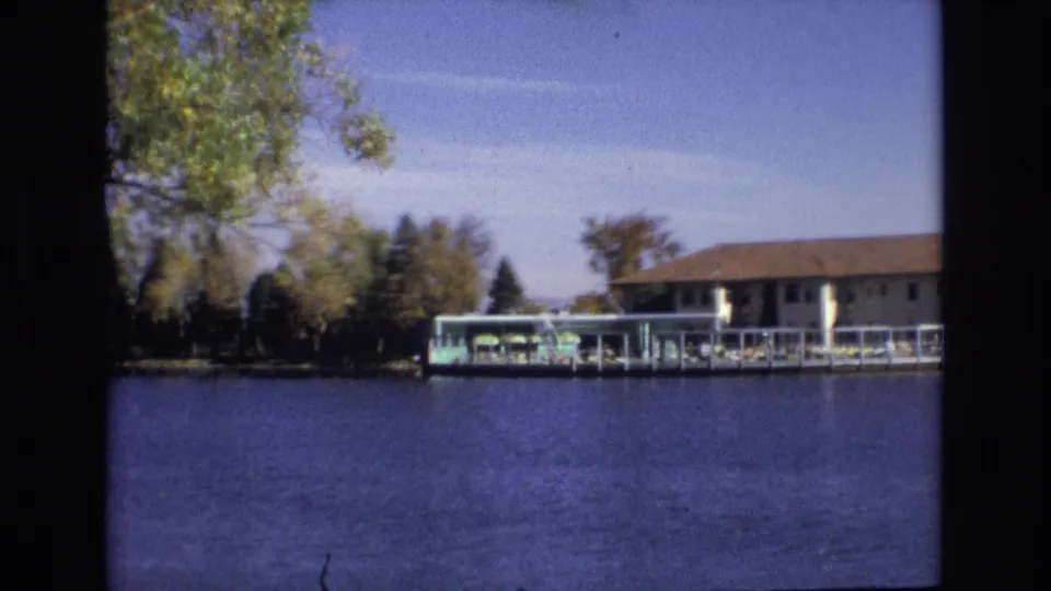 1974 Colorado Springs 8mm frame showing a long low lakeside building and covered dock across blue water, with shoreline trees and clear sky.