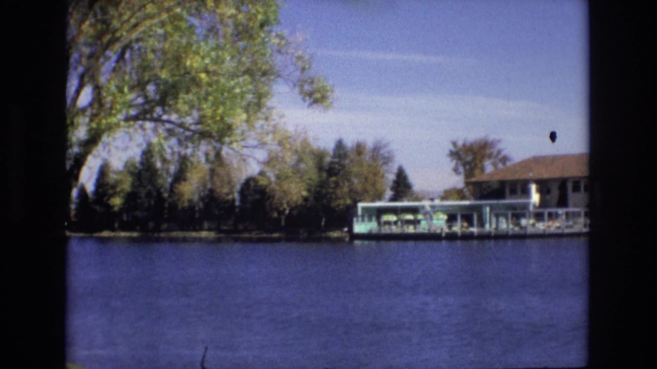 1974 Colorado Springs 8mm frame with a covered dock, low lakeside building, leafy trees, and blue water seen from across the lake.