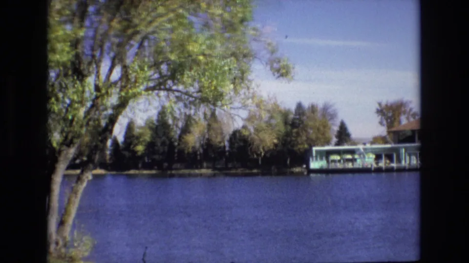 1974 Colorado Springs 8mm frame showing a large leafy tree at the lakeshore, blue water, and a covered dock partly visible across the lake.