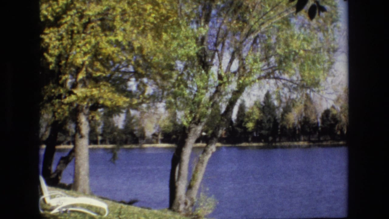 1974 Colorado Springs home-movie frame showing a calm pond behind leafy trees, with a white lounge chair partly visible at the lower left.