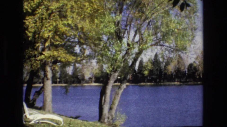 1974 Colorado Springs home-movie frame showing a calm pond behind leafy trees, with a white lounge chair partly visible at the lower left.