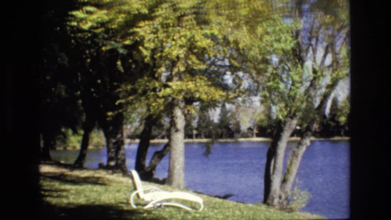 1974 Colorado Springs park scene with a white lounge chair on a grassy bank, mature trees overhead, and blue pond water beyond.