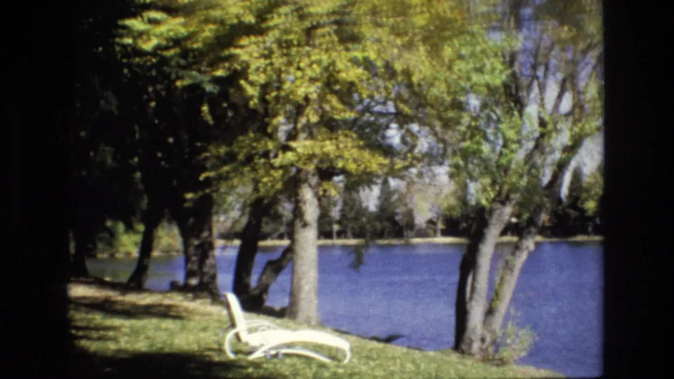 1974 Colorado Springs park scene with a white lounge chair on a grassy bank, mature trees overhead, and blue pond water beyond.