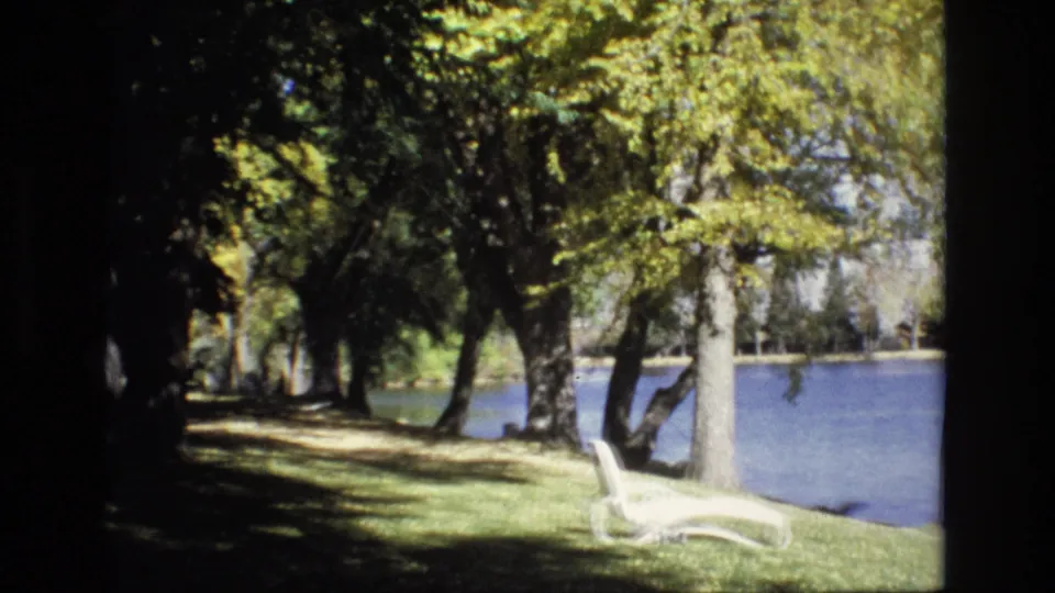 Handheld 8mm view of a shaded Colorado Springs pond bank in 1974, with trees framing blue water and a white lounge chair near the grass edge.