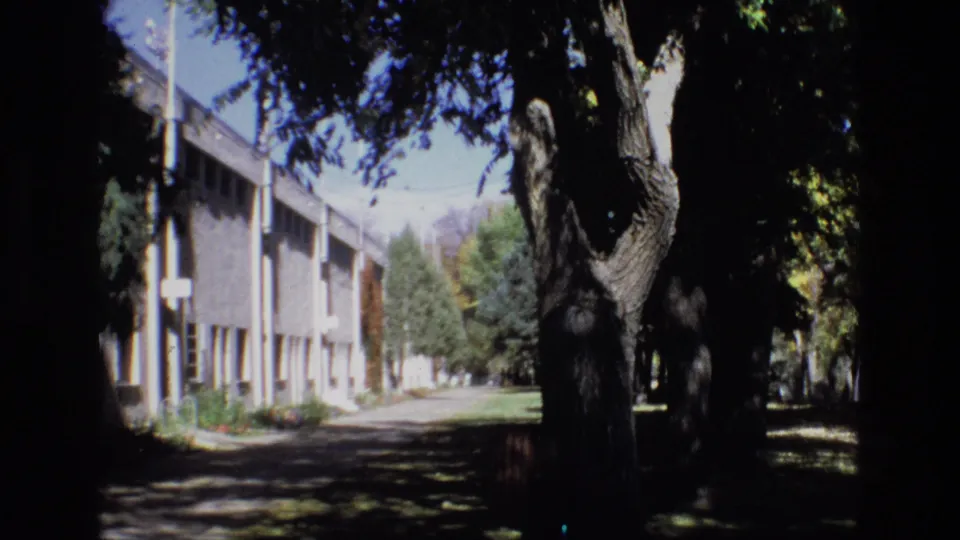 1974 Colorado Springs 8mm still showing a low-rise institutional-looking building with vertical columns beside a shaded walkway and mature trees.