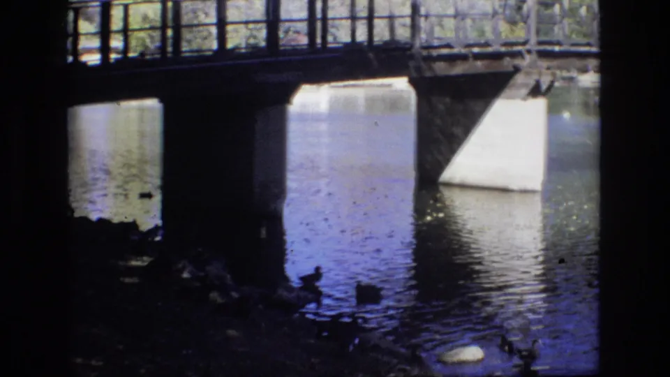 1974 Colorado Springs frame showing a bridge over still water, concrete piers, reflections, and ducks moving through the shaded foreground.