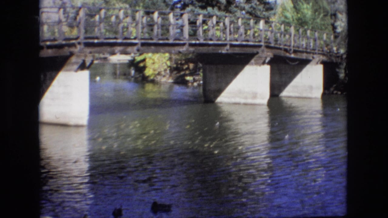1974 Colorado Springs 8mm frame showing rippling river water beneath a low bridge with wooden railings, concrete supports, trees, and ducks near the bottom edge.