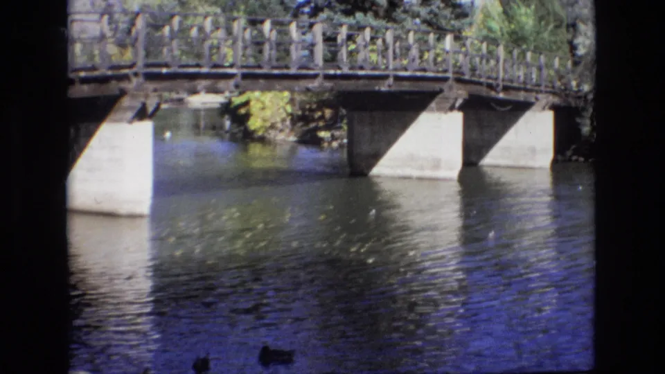 1974 Colorado Springs 8mm frame showing rippling river water beneath a low bridge with wooden railings, concrete supports, trees, and ducks near the bottom edge.