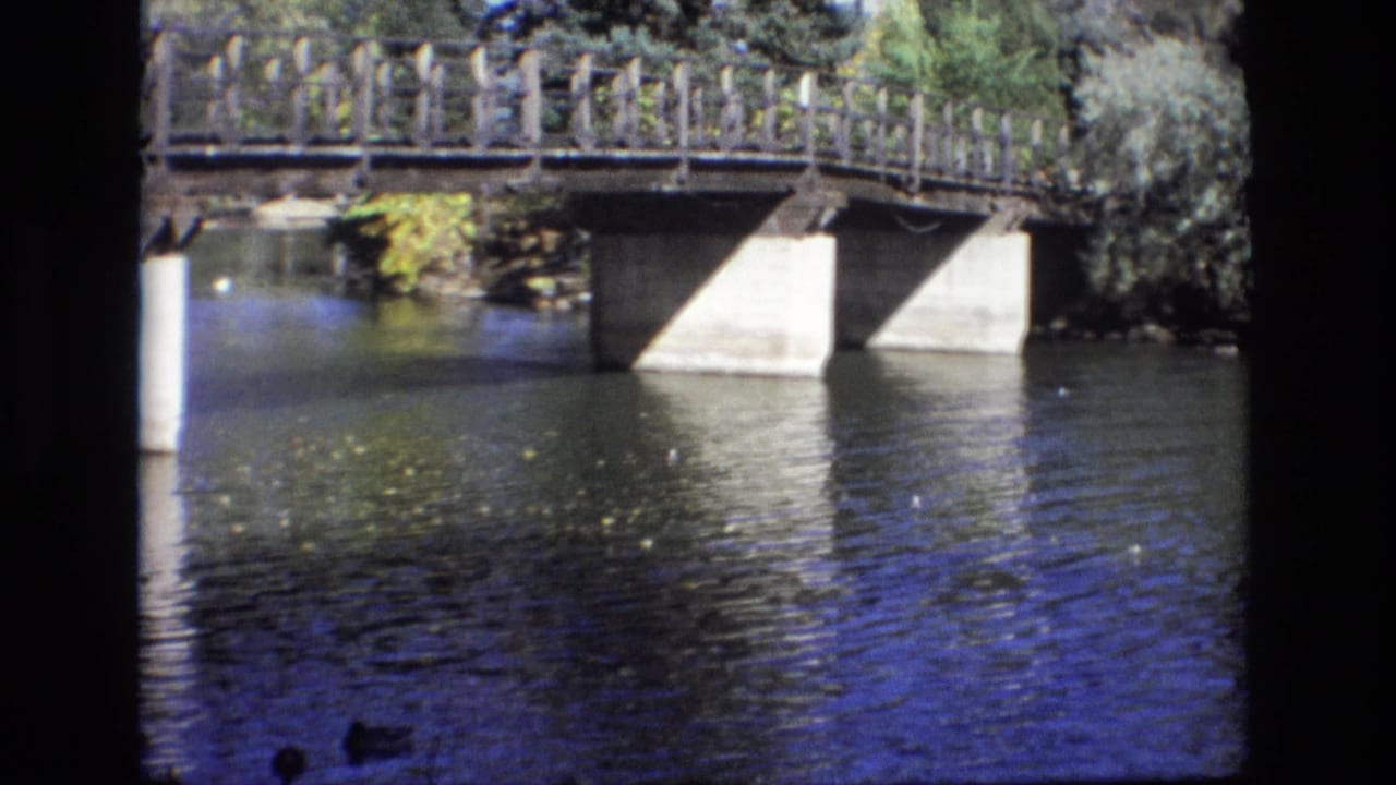 1974 Colorado Springs 8mm frame with a wooden-railed bridge over reflective river water, concrete piers, leafy banks, and ducks barely visible near the lower edge.