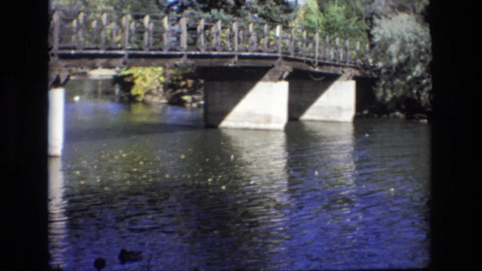 1974 Colorado Springs 8mm frame with a wooden-railed bridge over reflective river water, concrete piers, leafy banks, and ducks barely visible near the lower edge.