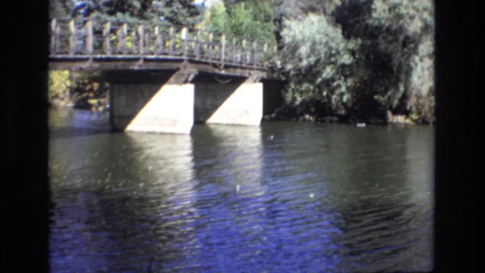 1974 Colorado Springs 8mm frame showing the bridge shifted right in view, with concrete supports, wooden railings, calm rippling water, reflections, and heavy foliage along the bank.
