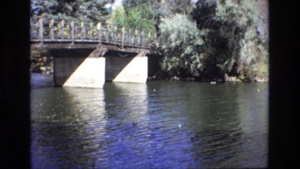 1974 Colorado Springs 8mm frame showing a wooden bridge over a river, with rippling water, leafy trees, and dark film borders at the edges.