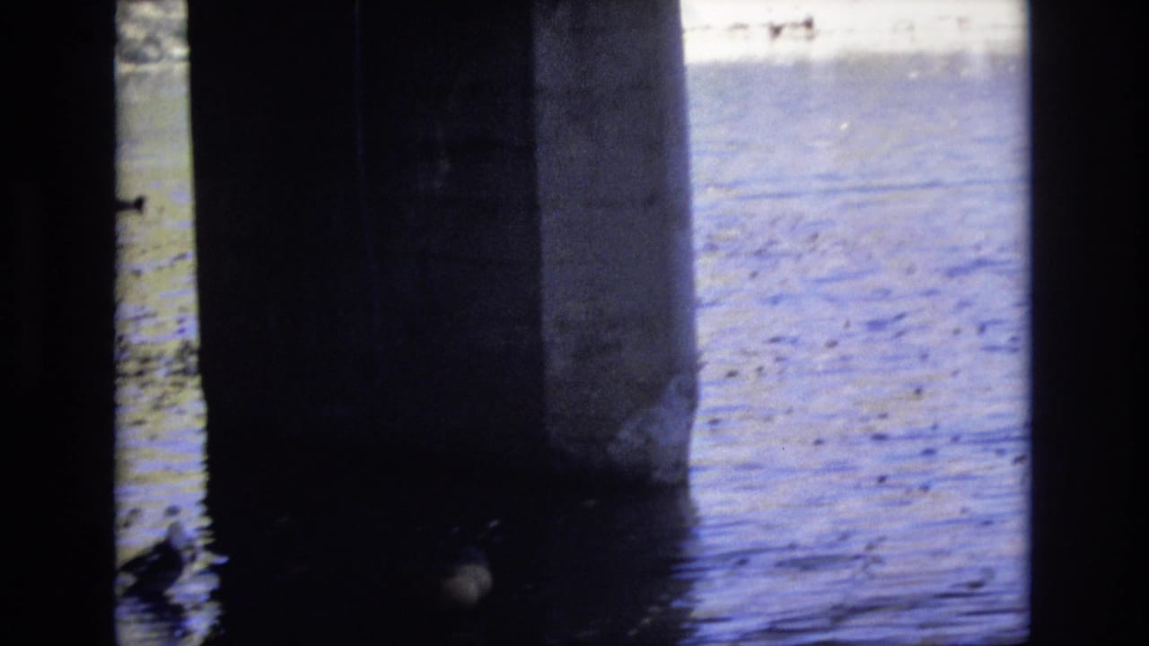 1974 Colorado Springs home-movie frame from beneath a concrete bridge, showing dark pillars, rippling blue water, and a duck near the shaded support.