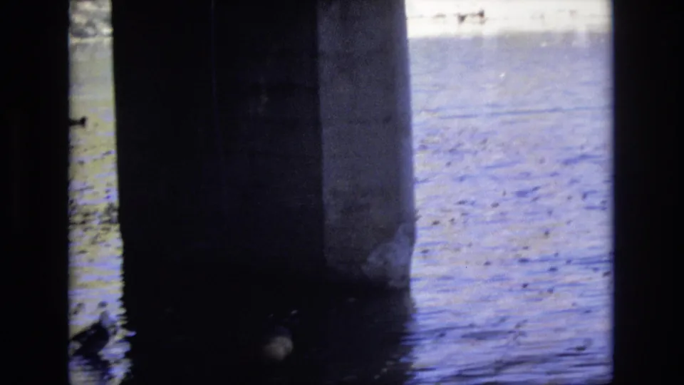 1974 Colorado Springs home-movie frame from beneath a concrete bridge, showing dark pillars, rippling blue water, and a duck near the shaded support.