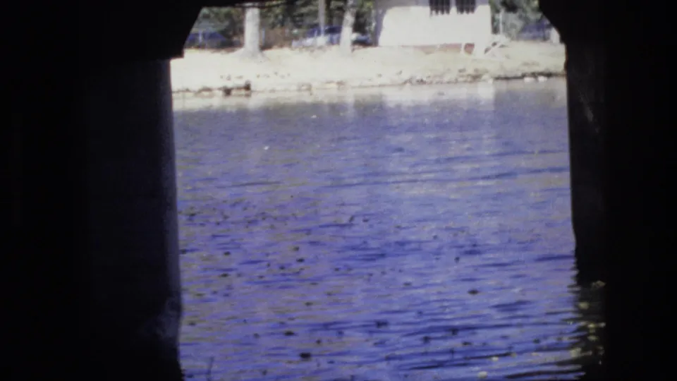 A water-level 8mm view beneath a bridge in Colorado Springs, with dark concrete supports framing rippling water and a light shoreline in the distance.
