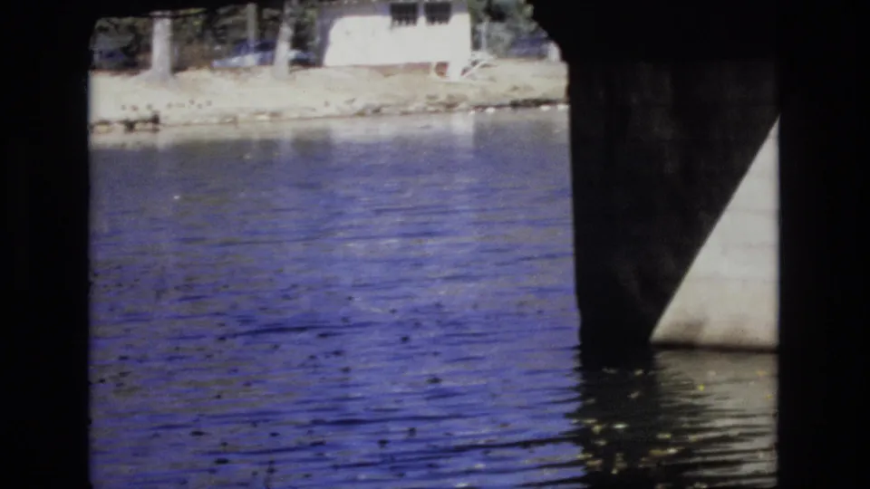 1974 home-movie frame showing calm water beneath a bridge, with a pale shoreline, trees, and a distant house or building across the water.
