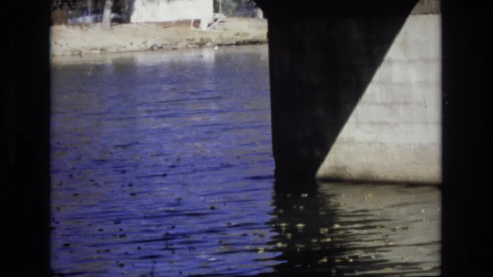 A final 8mm frame from under the bridge, showing blue-tinted rippling water, a dark support, and a pale concrete bridge edge or embankment at the right.