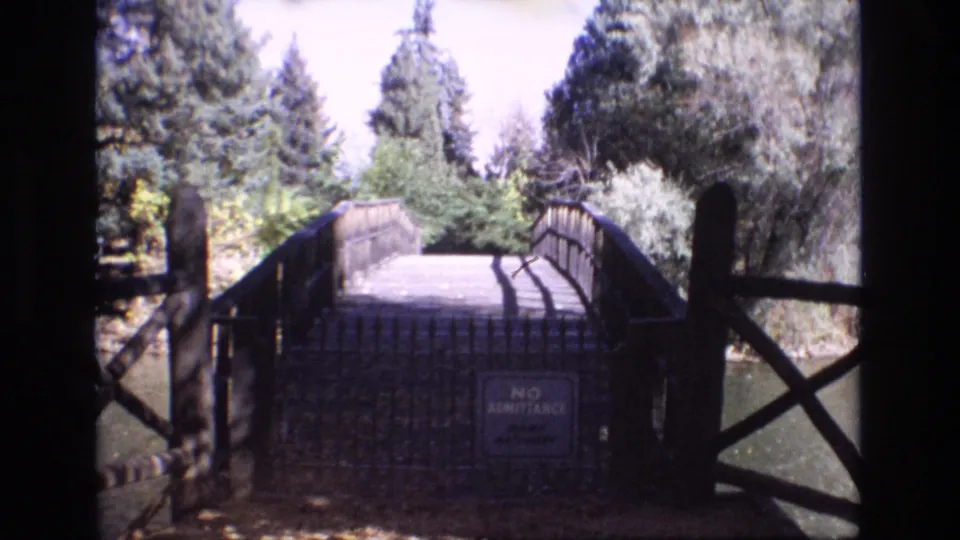 1974 Colorado Springs home-movie frame showing a small wooden bridge or walkway with railings, dense trees, and a visible no-admittance sign.