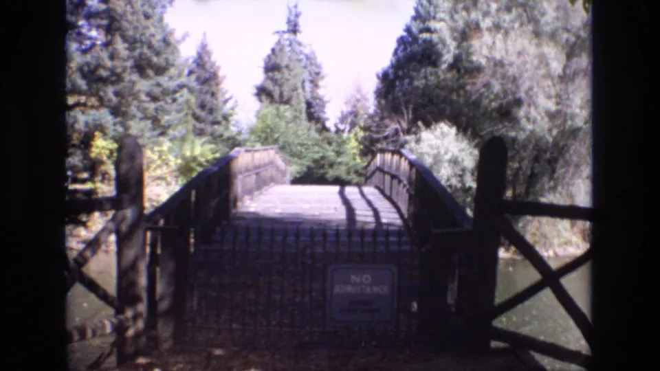 1974 Colorado Springs home-movie frame showing a rustic wooden bridge over calm water, framed by dark railings, trees, and a visible no-admittance sign.