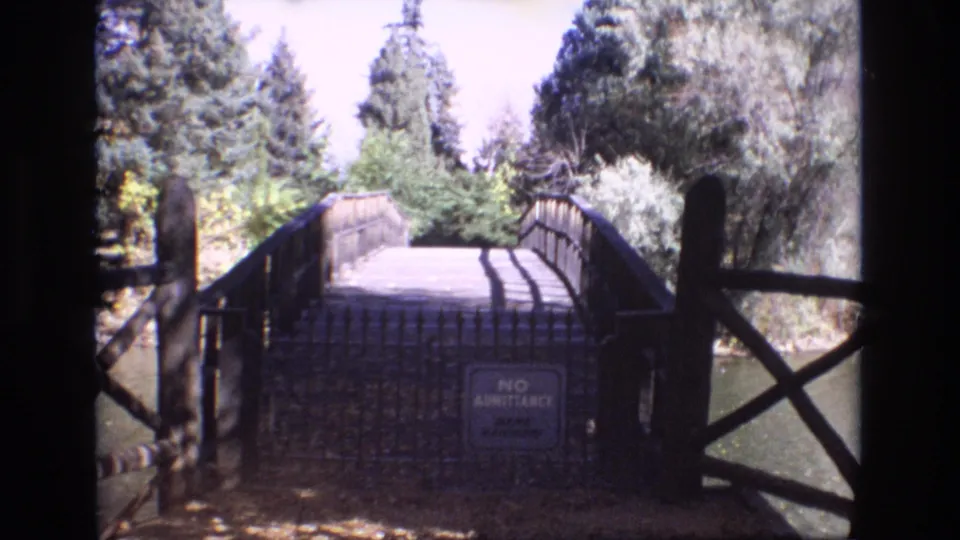 1974 Colorado Springs 8mm frame of a rustic bridge spanning calm water, with dark railings close to the camera and a no-admittance sign on the bridge approach.