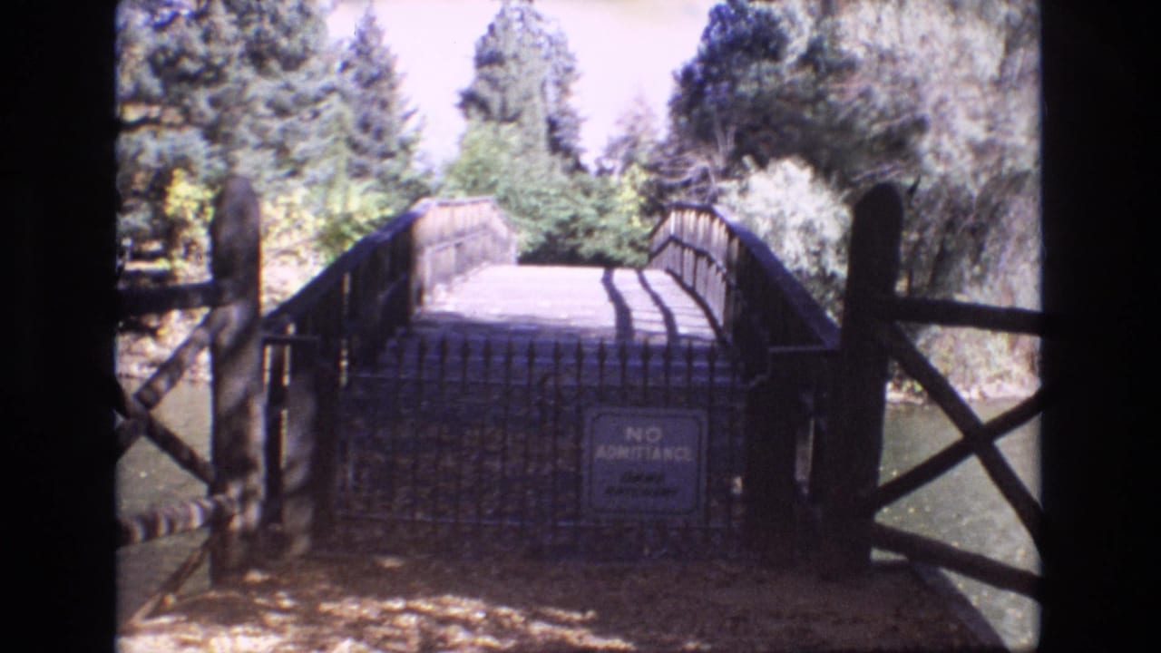 1974 Colorado Springs 8mm frame showing a wooden pedestrian bridge entrance, metal gate, no admittance sign, pond edge, and surrounding trees.