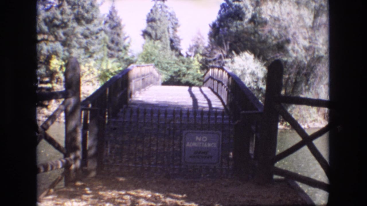 1974 Colorado Springs park view of a gated wooden bridge crossing near water, framed by dark railings and leafy trees.