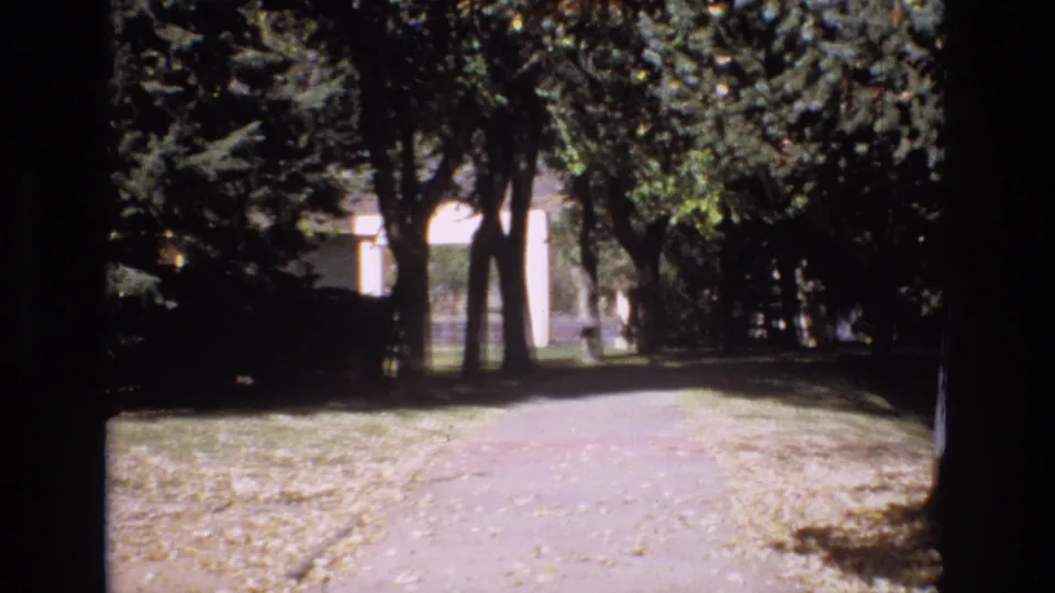 1974 Colorado Springs 8mm frame looking down a shaded park path lined with trees, grass, fallen leaves, and a distant light-colored building.