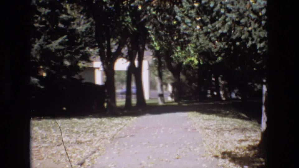 1974 Colorado Springs park path in soft 8mm color, with trees casting shade across the walkway and a light-colored building visible beyond.