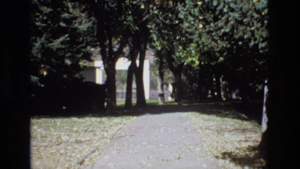 1974 Colorado Springs 8mm frame showing the walkway from a slightly shifted home-movie angle, with leaves on the ground and a pale building filtered through trees.